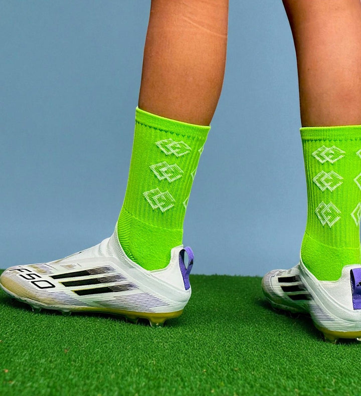 Player wearing lime green C3 grip socks with white soccer cleats on green turf