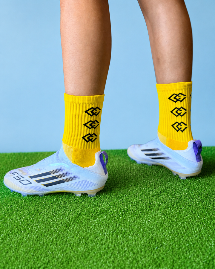 Player wearing yellow C3 grip socks with white soccer cleats on green turf
