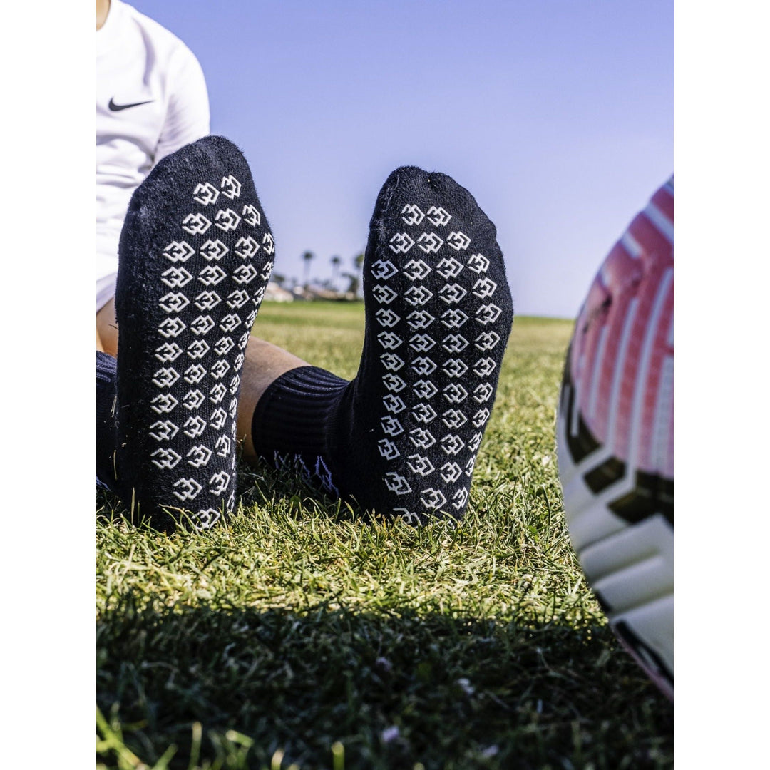 Soles of black C3 grip socks showing the white C3 logo grip pattern beside a soccer ball on grass