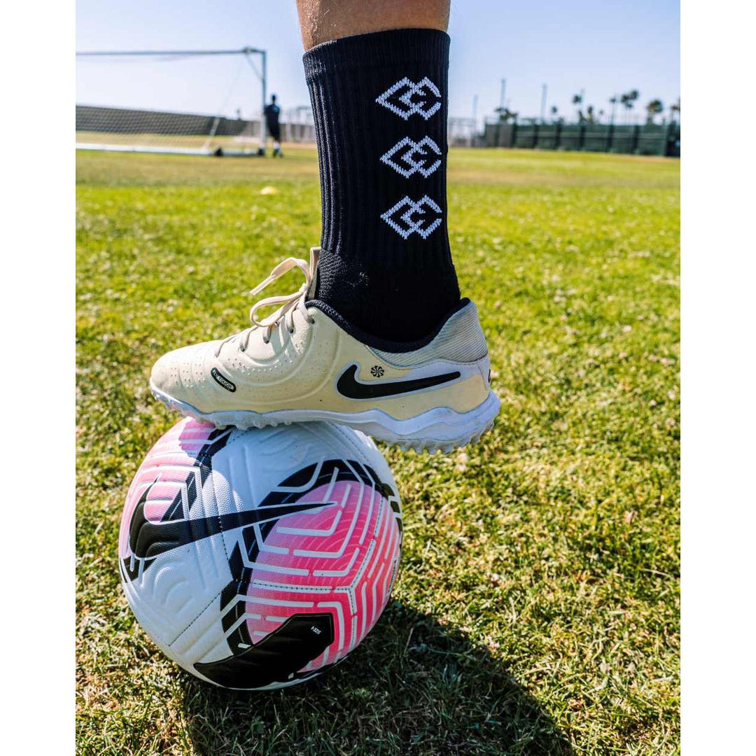 Player wearing black C3 grip socks and white soccer cleats with one foot on a soccer ball