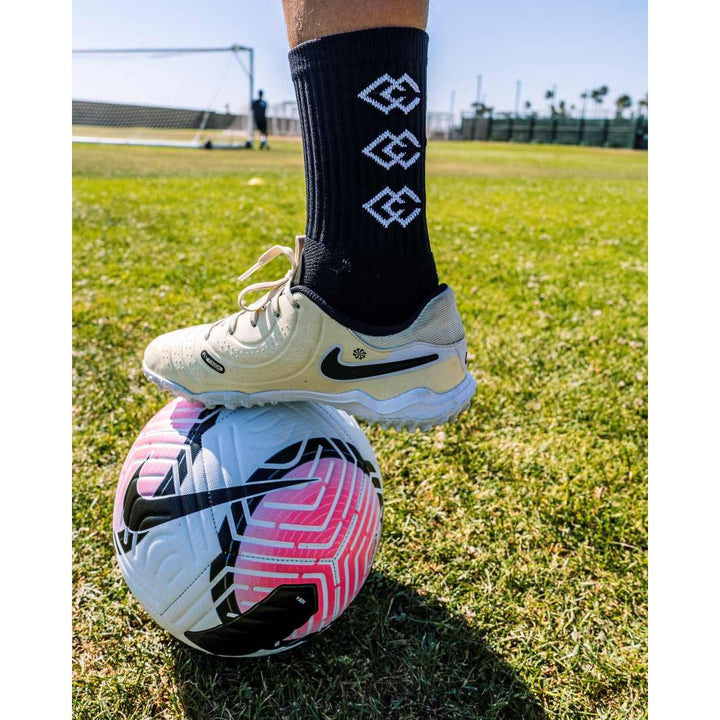 Player wearing black C3 grip socks and white soccer cleats with one foot on a soccer ball
